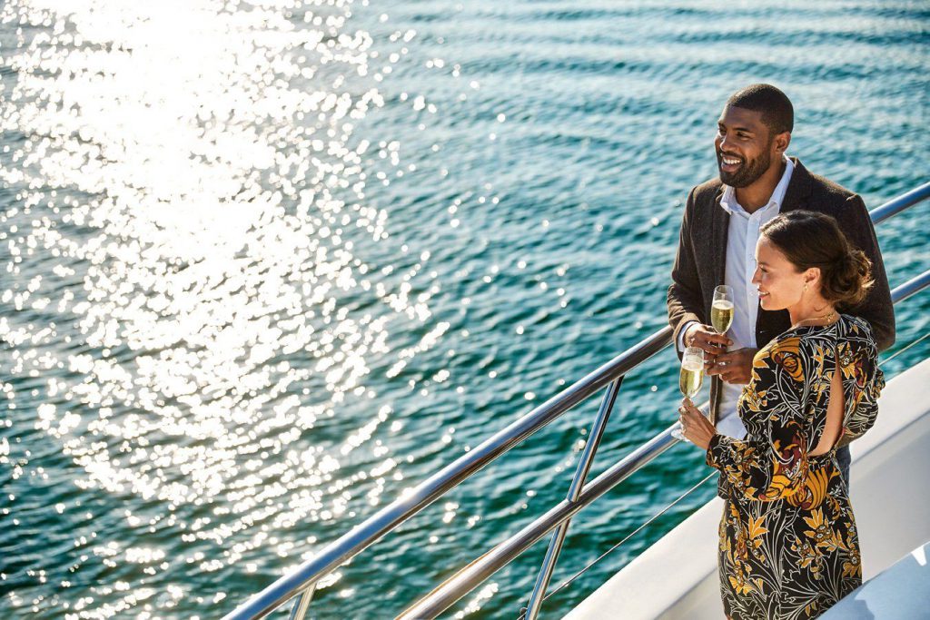 Couple stands on deck of boat holding champagne glasses with water in background.