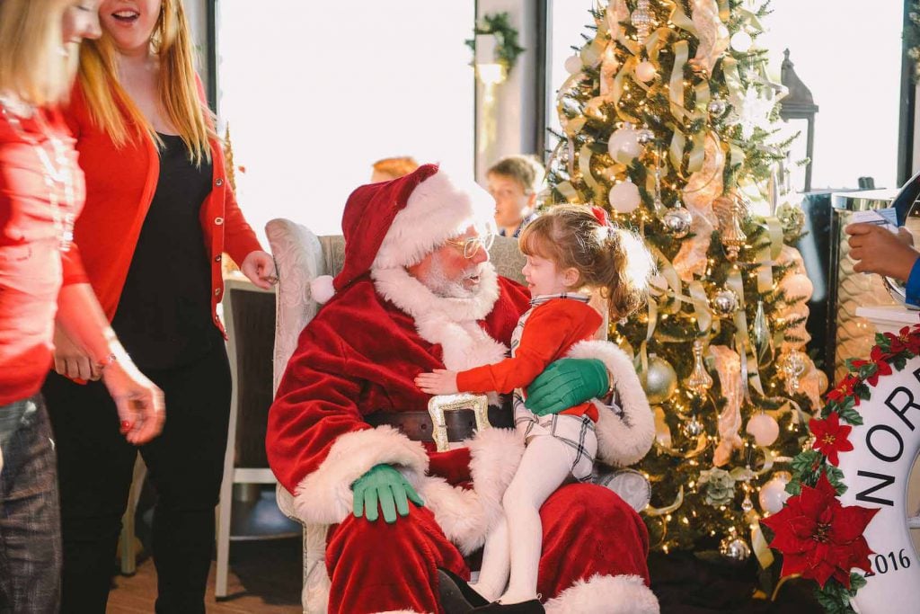 Santa with a little girl sitting on his lap and a Christmas tree in background.