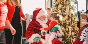 Santa with a little girl sitting on his lap and a Christmas tree in background.