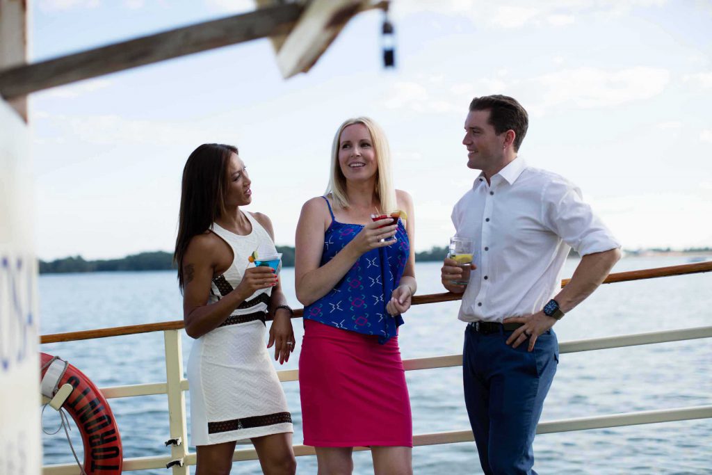Three people leaning on outer deck boat railing holding drinks.