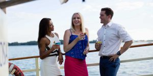 Tres personas apoyadas en la barandilla de la cubierta exterior del barco sosteniendo bebidas.