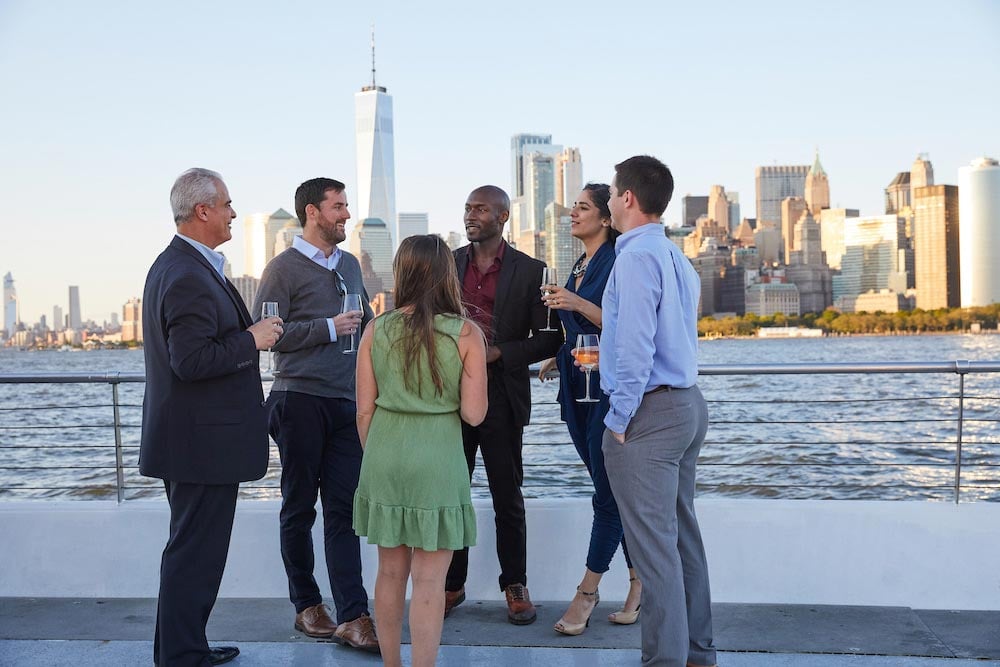 Groep mensen op bootdek met skyline van New York City op achtergrond.