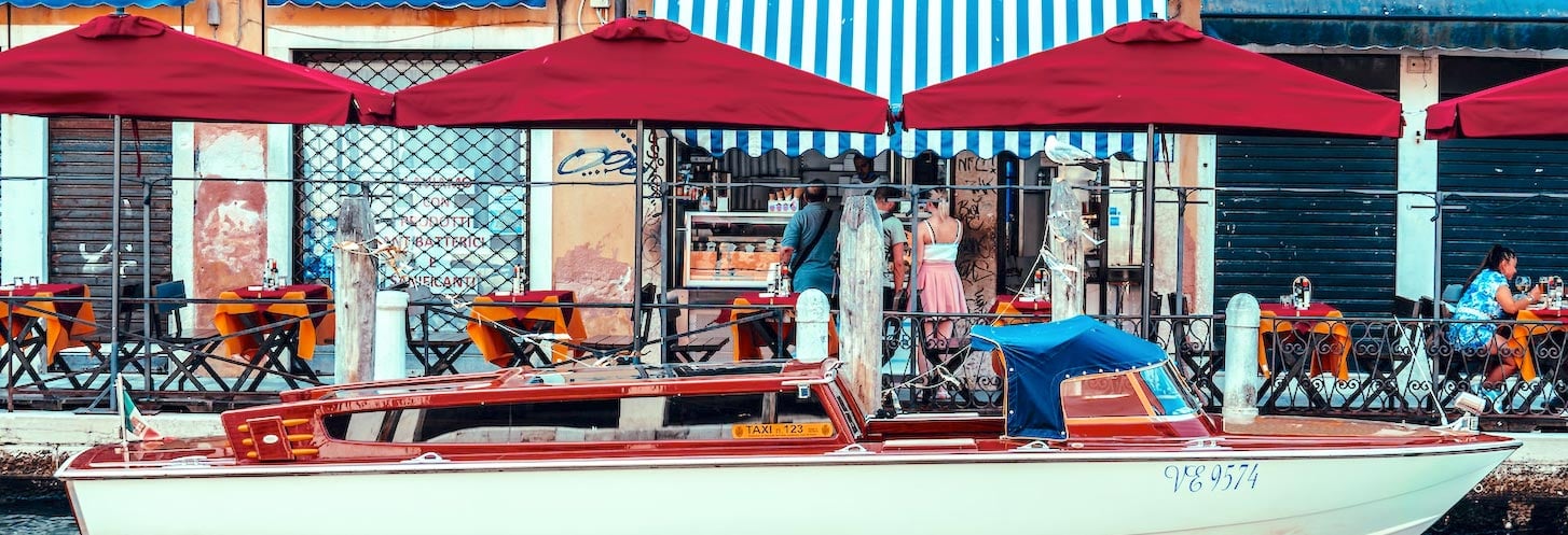 Venice Italy restaurant with boat moored in front and people at tables.